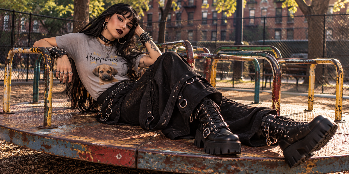 A person lounging on a rusty playground merry-go-round wearing a gray graphic t-shirt with a dog print, black lace-up cargo pants, and black combat boots in an outdoor park setting.